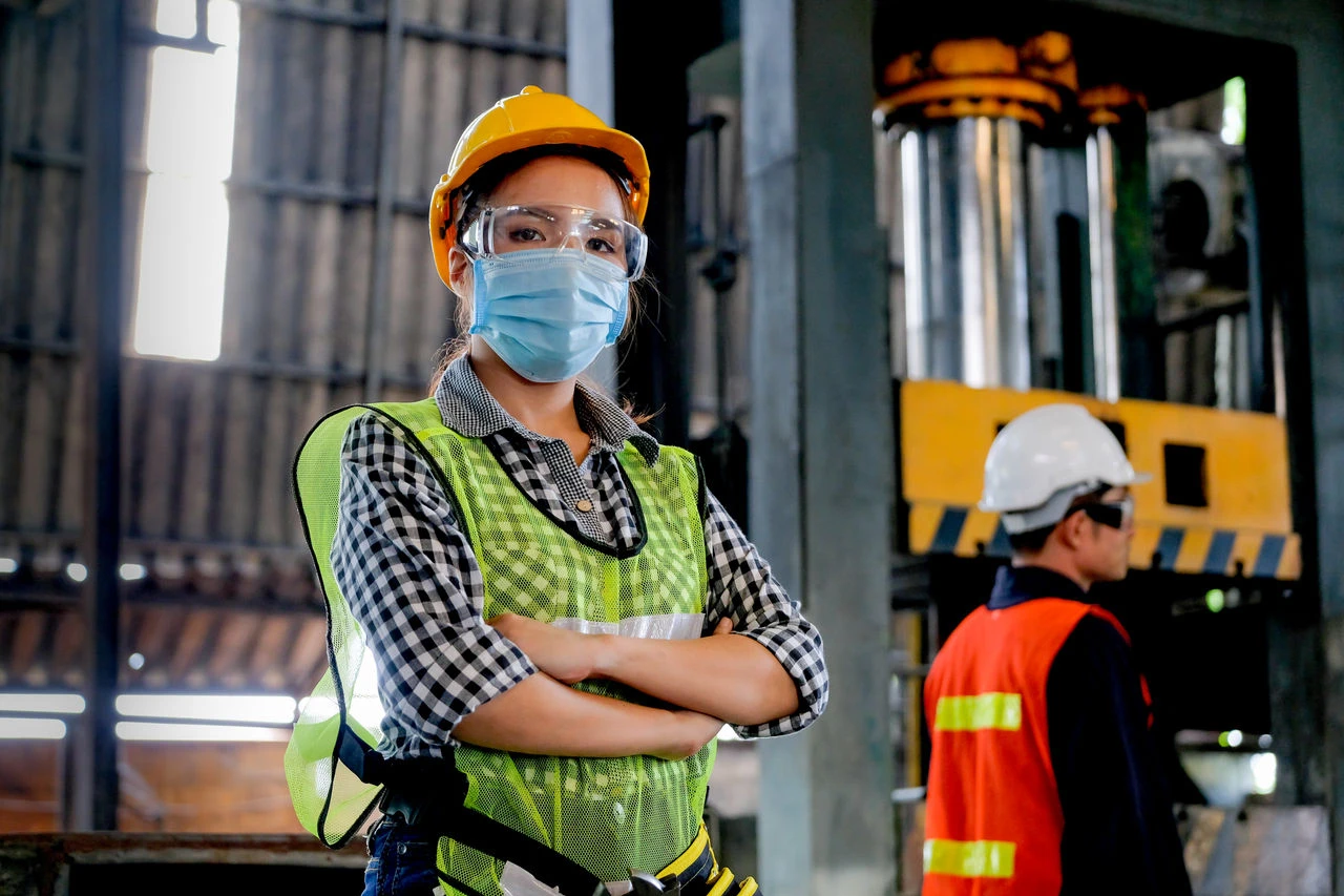 A woman wearing a hard hat, safety glasses, mask, and high-visibility vest in an industrial workplace